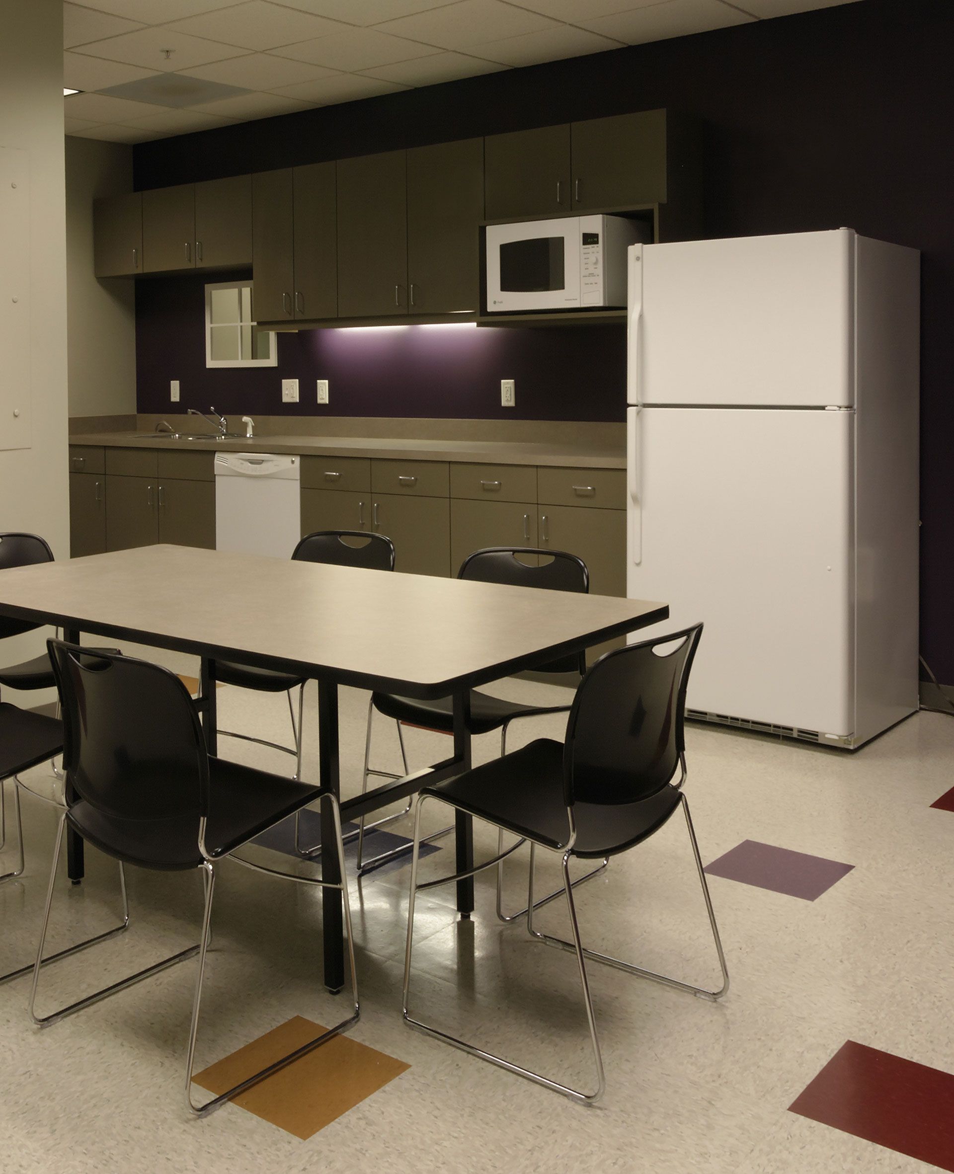 A breakroom with a table, chairs, tan cabinets, a white refrigerator, a microwave, and a dishwasher against a purple wall.