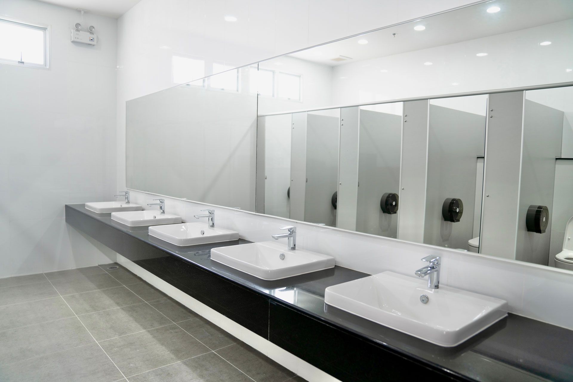 A row of white rectangular sinks on a black countertop in a modern, brightly lit public restroom.