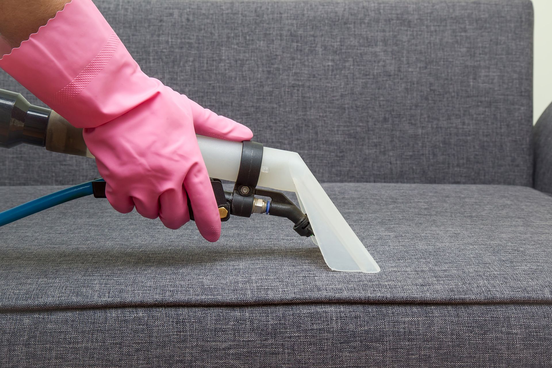A person wearing pink gloves is cleaning a couch with a vacuum cleaner.
