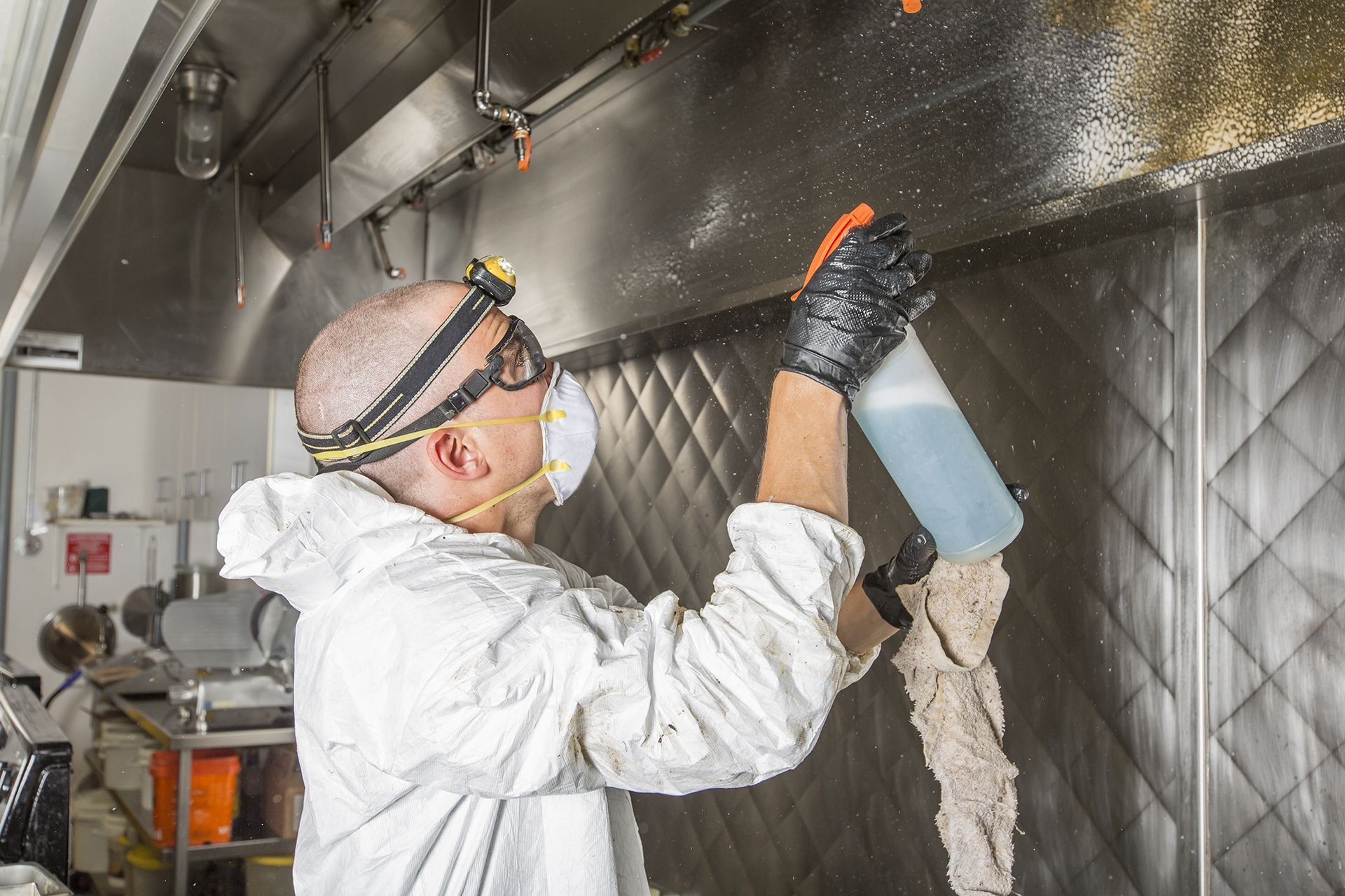 A man wearing a mask and gloves is cleaning a kitchen hood with a spray bottle.