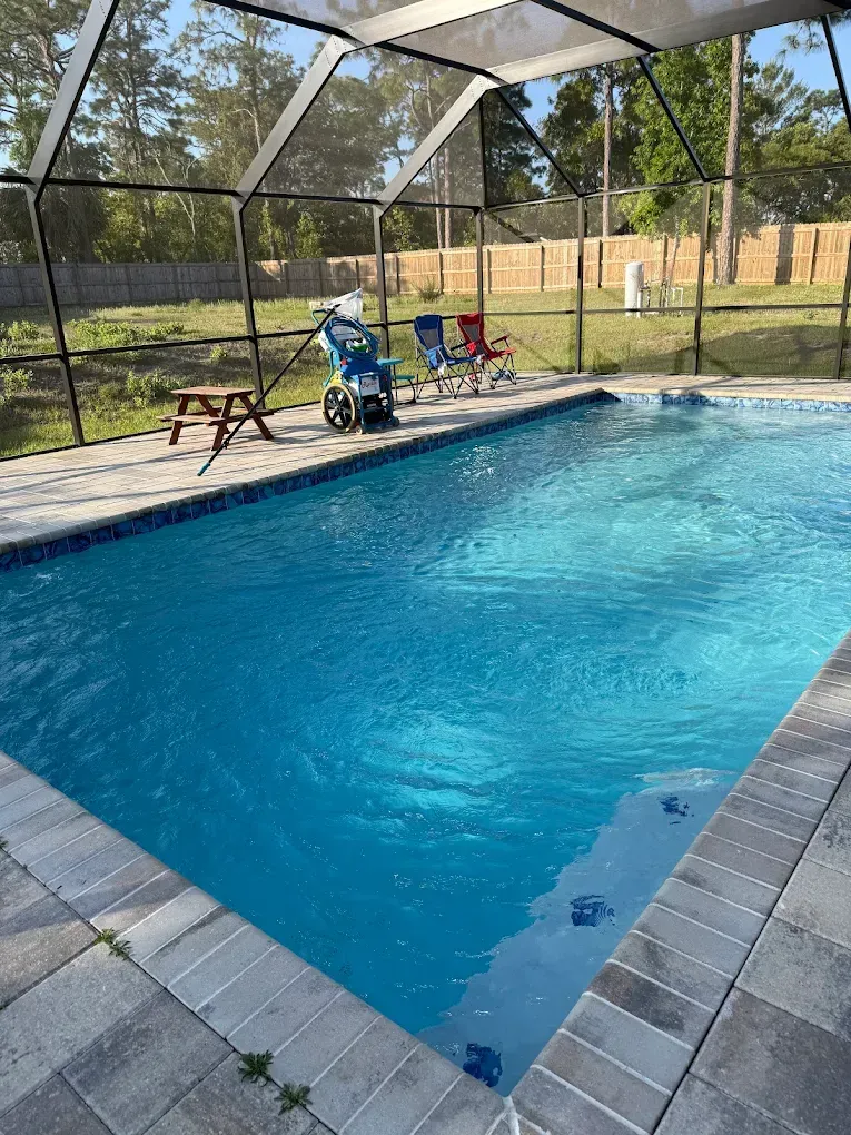 Swimming pool with blue water under a screened enclosure; a wheelchair and chairs are nearby.
