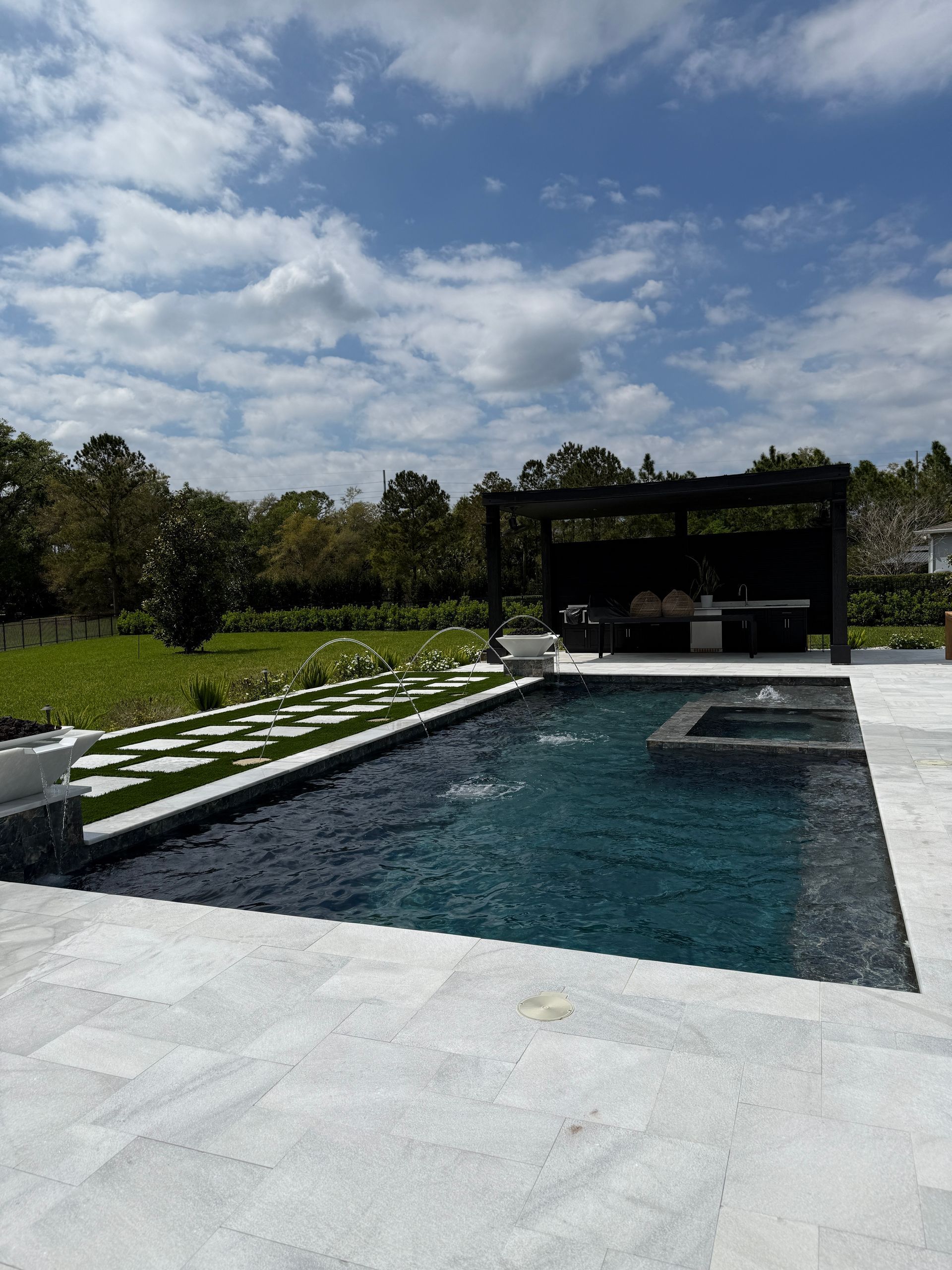 Swimming pool with a dark blue surface, surrounded by light gray stone patio. A pergola and green lawn are in the background.