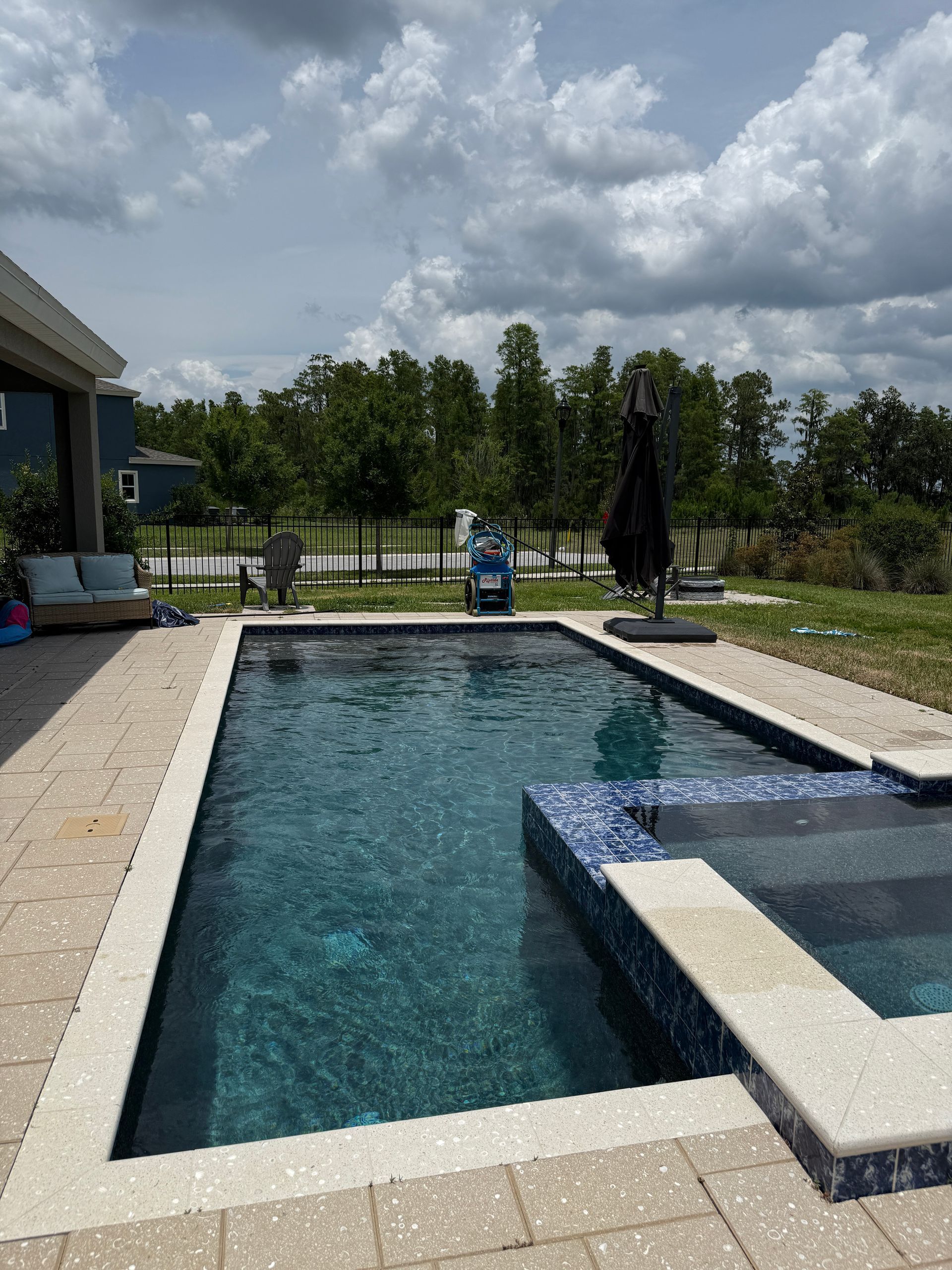 Rectangular pool with turquoise water, surrounded by tan patio, blue sky, and trees.