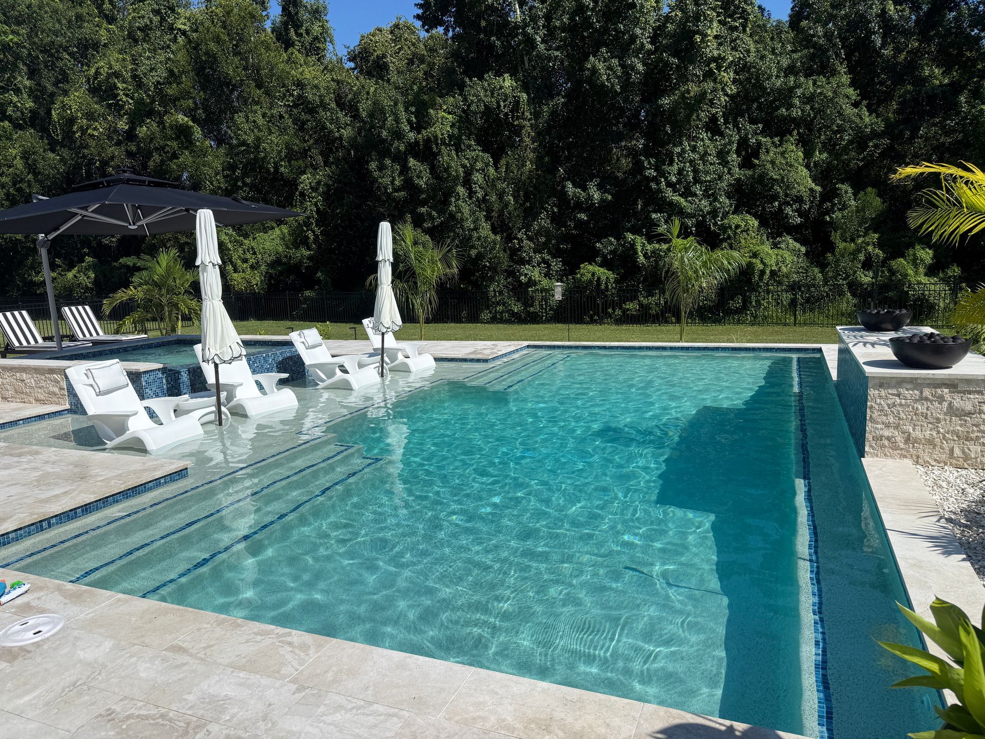 Pool with white lounge chairs, umbrellas, and lush greenery in the background on a sunny day.