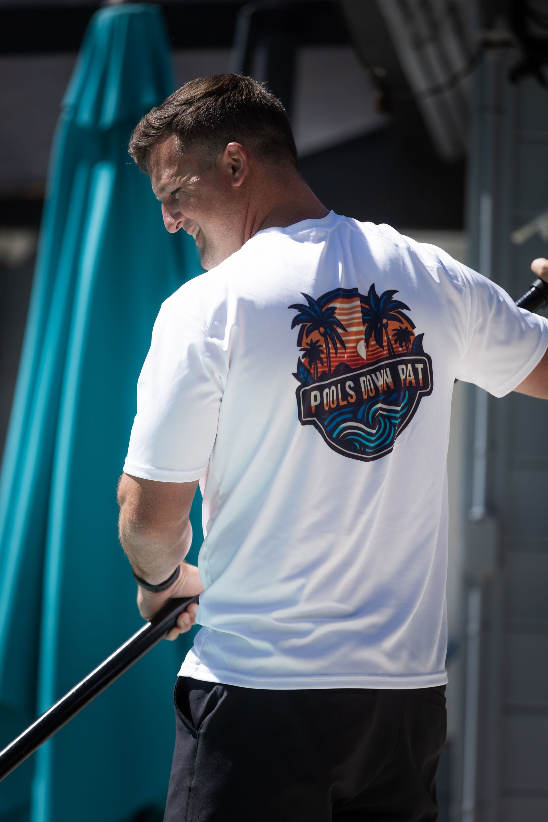 Man in white shirt with beach logo, holding a paddle, standing outdoors.