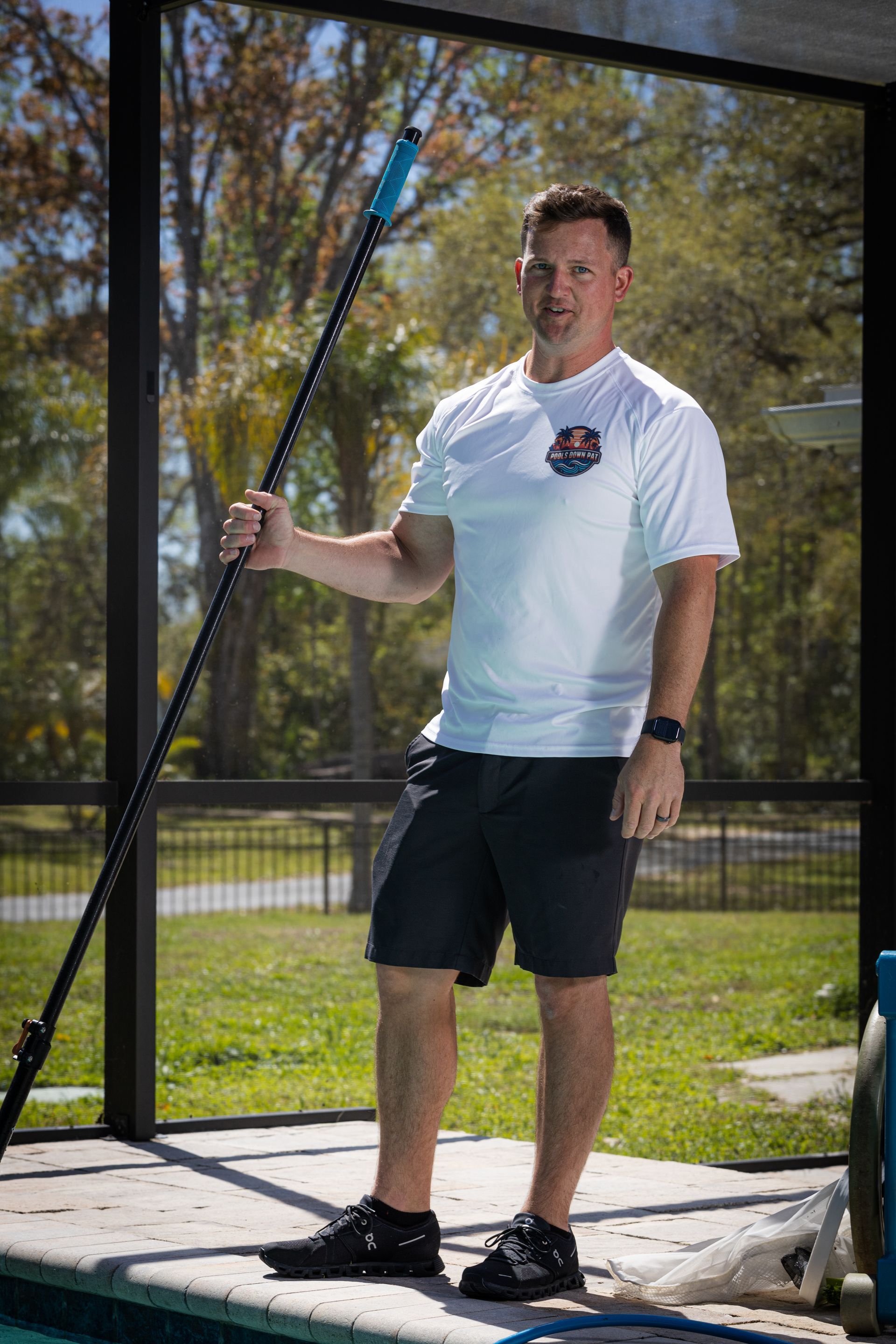 Man holding a long pool cleaning pole, standing near a pool, wearing a white shirt and black shorts.