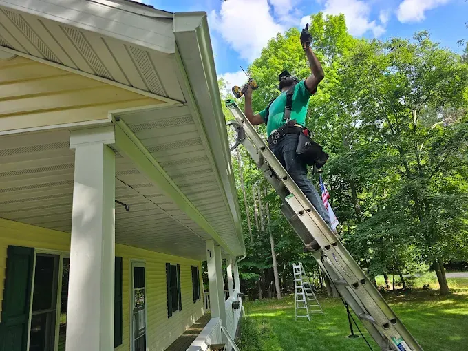 Person on ladder, installing siding on a house with green shutters and a white porch.