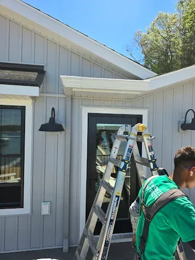 Man on ladder near a light gray house with a black door and lights.