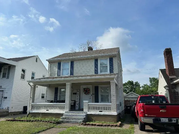 Two-story beige house with blue shutters and porch. Red truck parked on the side. Overcast sky.