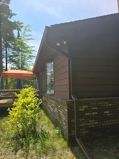 Brown house with brick base, windows, and gutter; green foliage in front, blue sky.