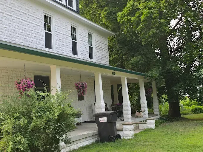 White house with a green roof and porch, surrounded by trees and green grass.