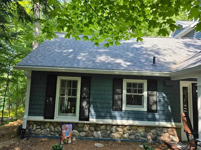 Blue house with white-framed windows and black shutters, rock foundation, nestled in trees.