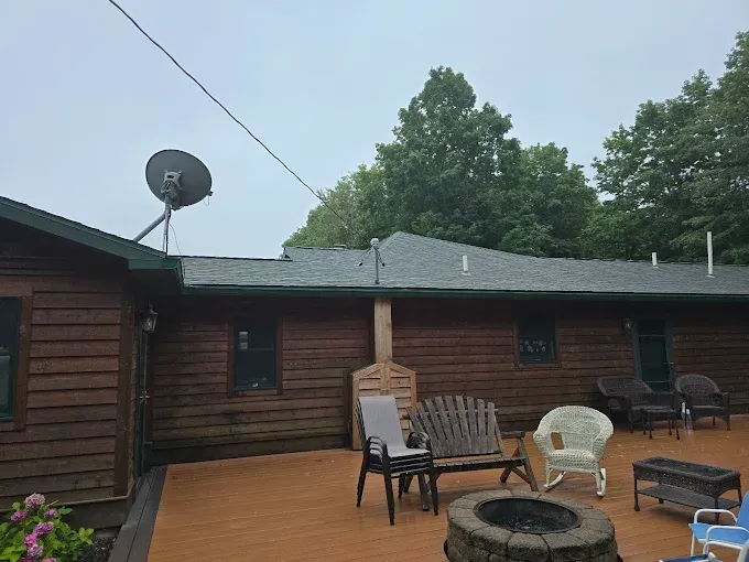 Brown cabin with a wooden deck, chairs, fire pit, and satellite dish under a cloudy sky.