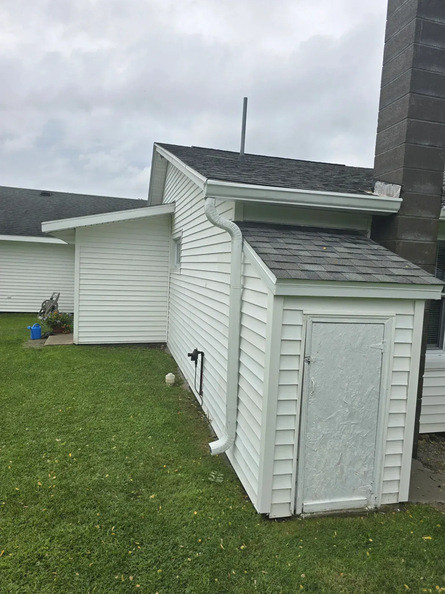 White building with a black shingled roof, next to a grassy yard and a cloudy sky.