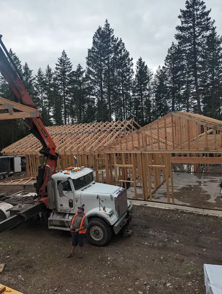 Construction site with a truck crane lifting lumber to frame a house. A worker stands nearby.