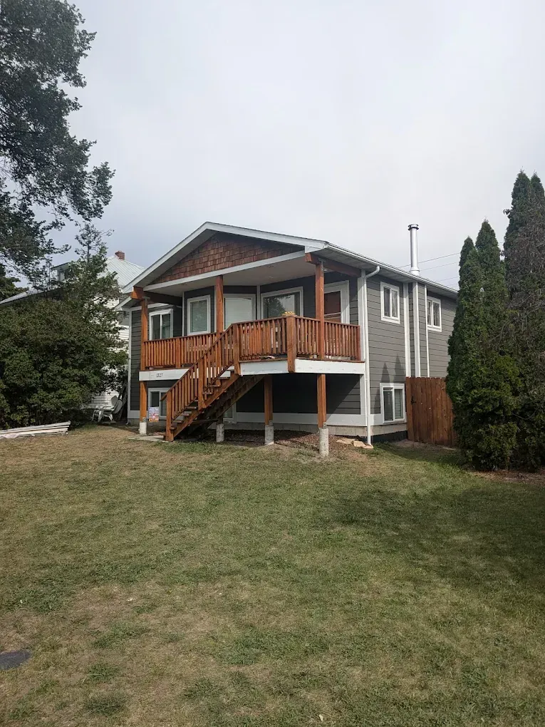 Two-story house with a wooden deck and stairs. The house has gray siding and a brown roof. A lawn is in front.