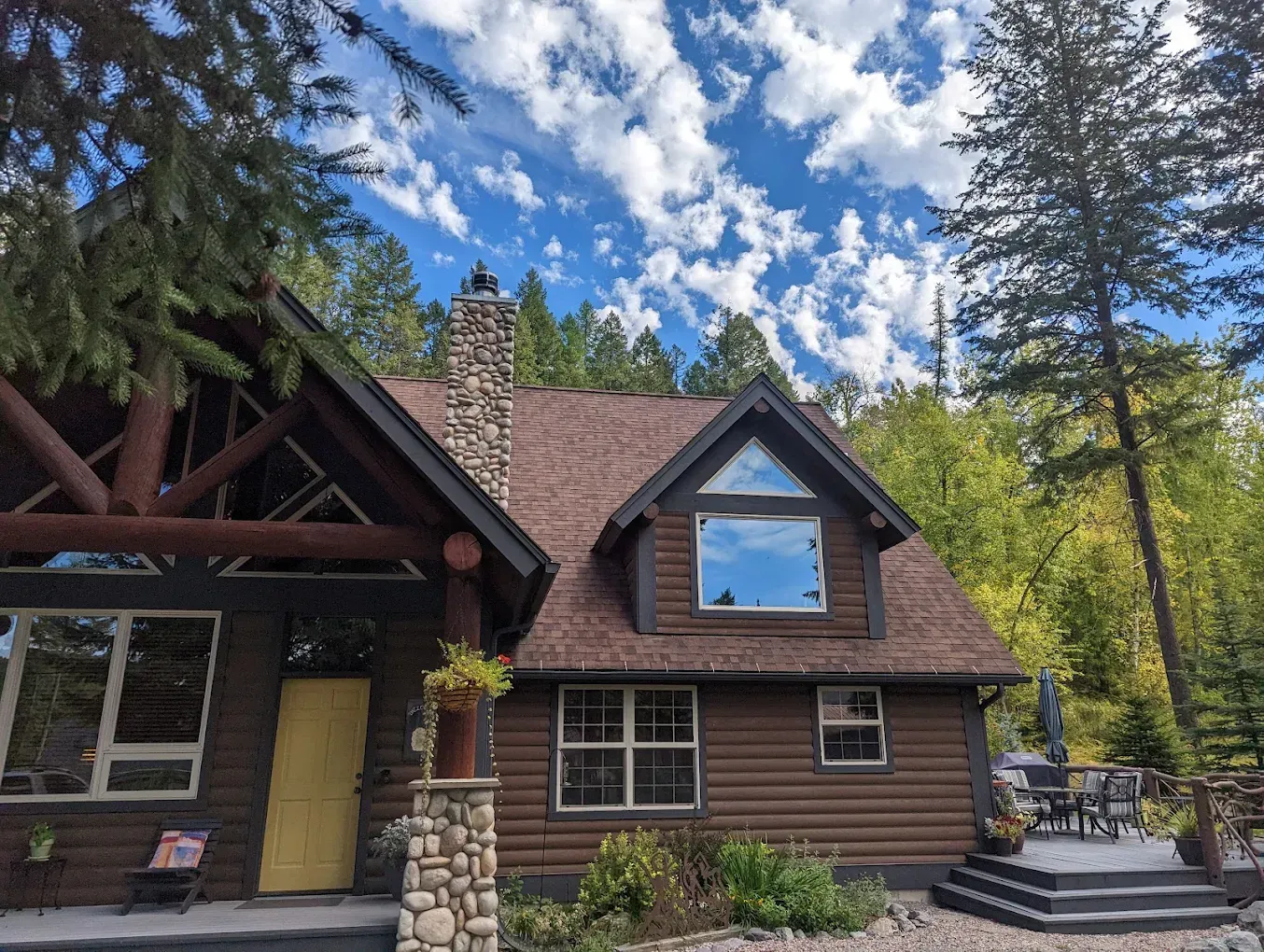 Brown log cabin with stone chimney, yellow door, under a blue sky with clouds.