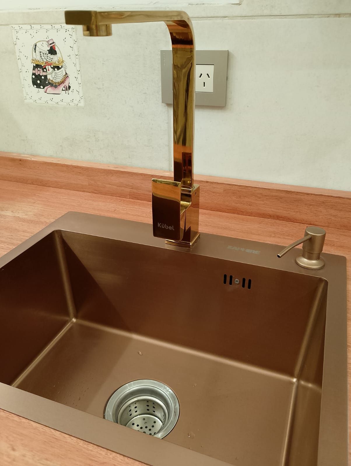 Grifo dorado sobre un fregadero de cocina de color cobre, con un dispensador de jabón a la derecha.