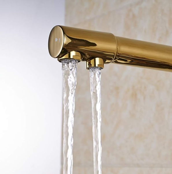 Grifo de color dorado con dos caños que vierten agua sobre un fondo de azulejos beige.