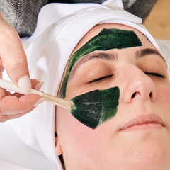 Woman receiving a facial treatment; lying down with towel on head, hands on face.