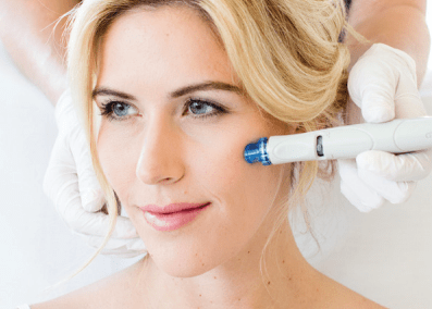 Woman receiving a facial treatment with a blue and white handheld device; white gloved hands, light skin, blonde hair.
