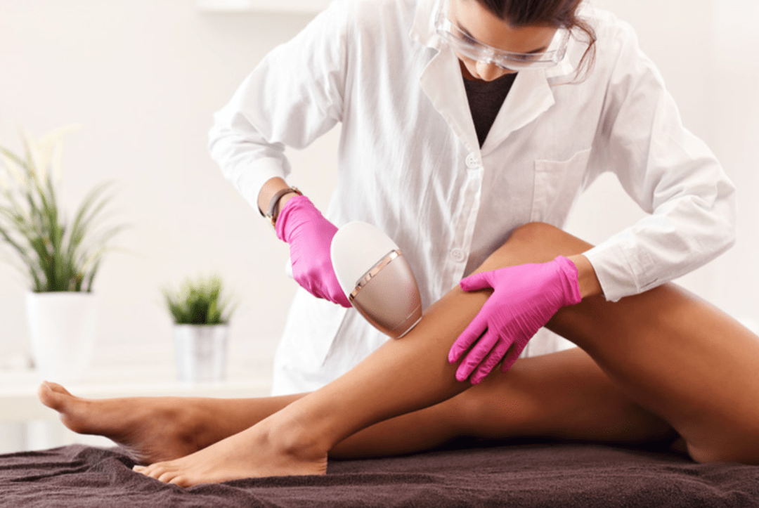 Person in white coat and pink gloves using a laser hair removal device on a leg in a bright room.