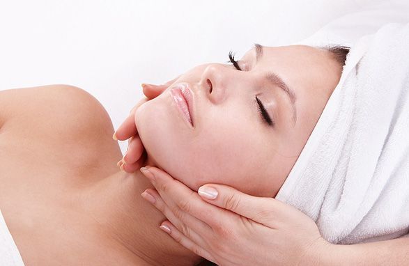 Woman receiving a facial treatment; lying down with towel on head, hands on face.