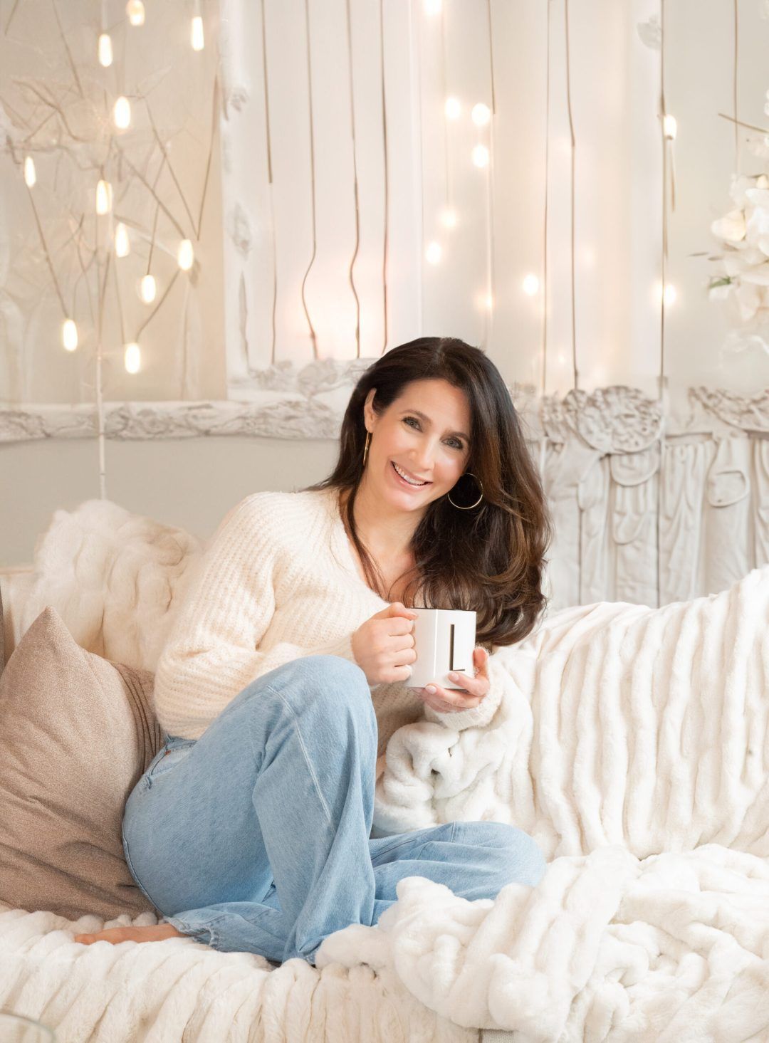 Woman sitting on a white couch, holding a mug, smiling. Decorated room with soft lighting.