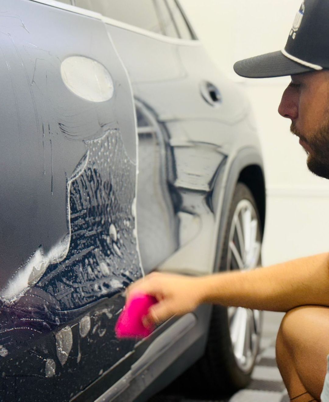 A man in a hat is cleaning a car with a pink sponge