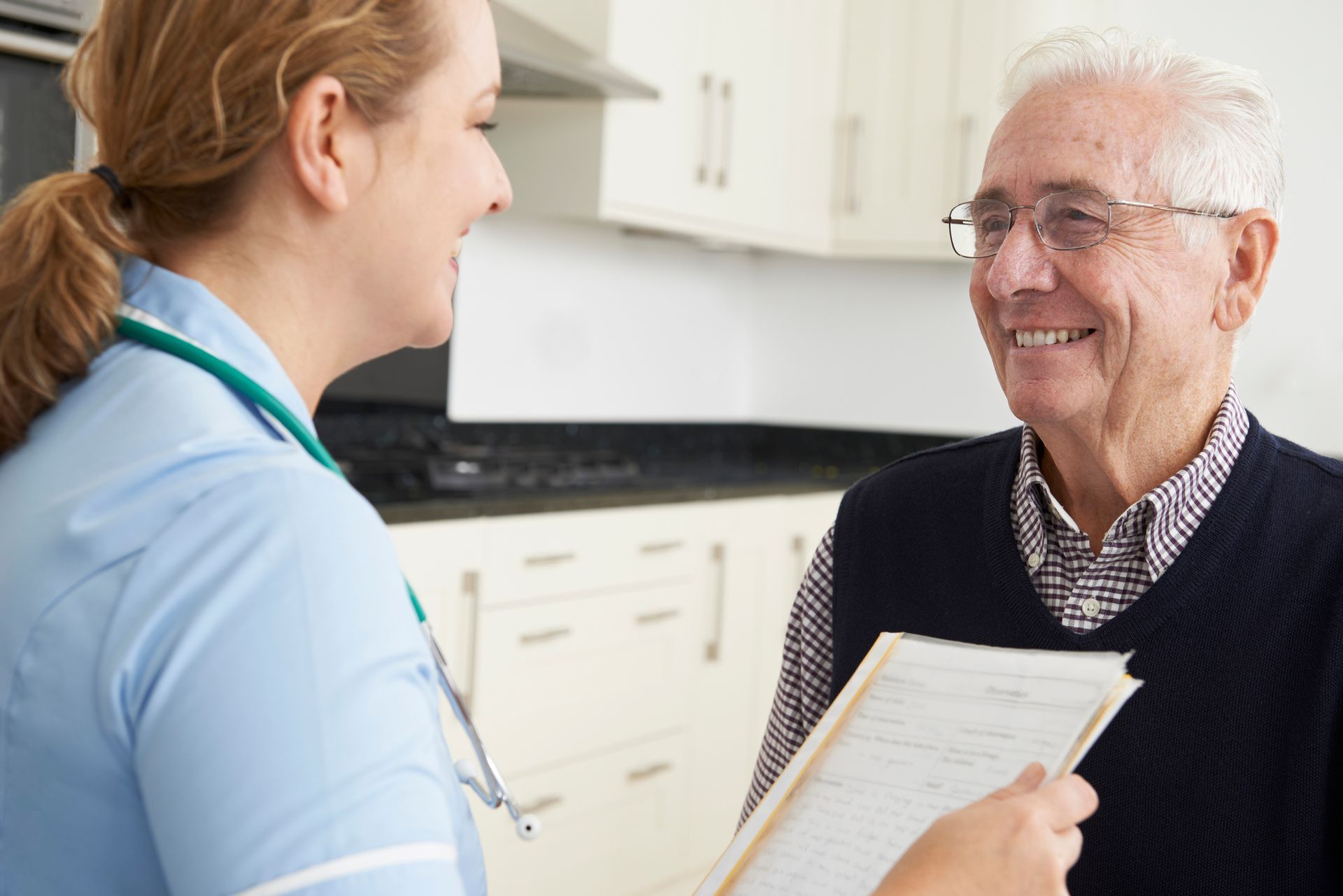 Nurse in blue scrubs smiles at senior man, holding clipboard, in a kitchen.