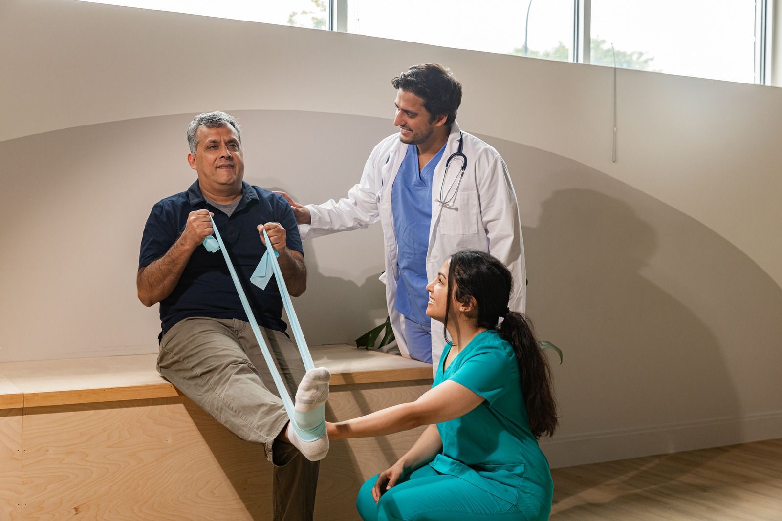 Man doing physical therapy with blue resistance band, guided by two medical professionals.
