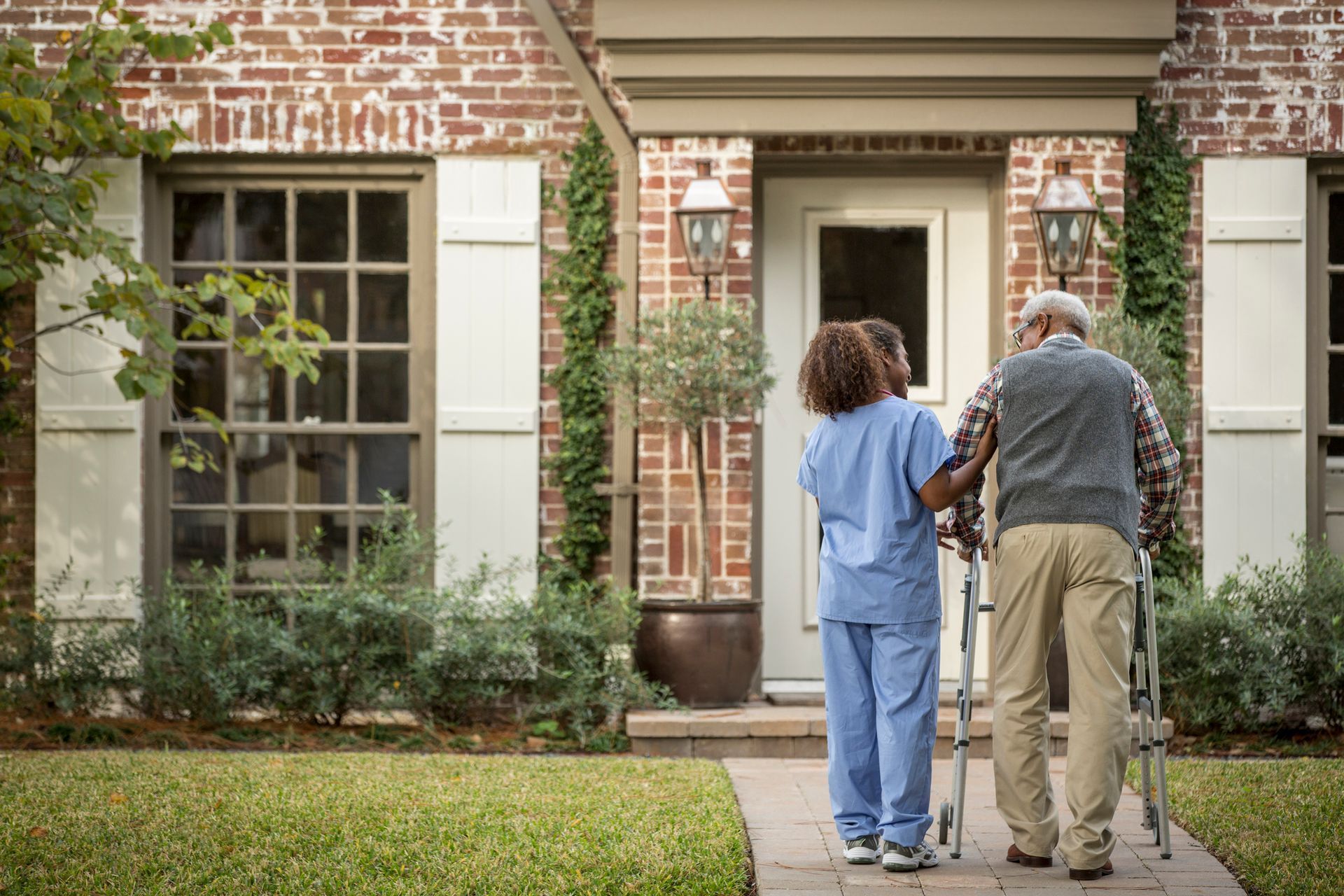 Caregiver assists a person with a walker towards a brick home.