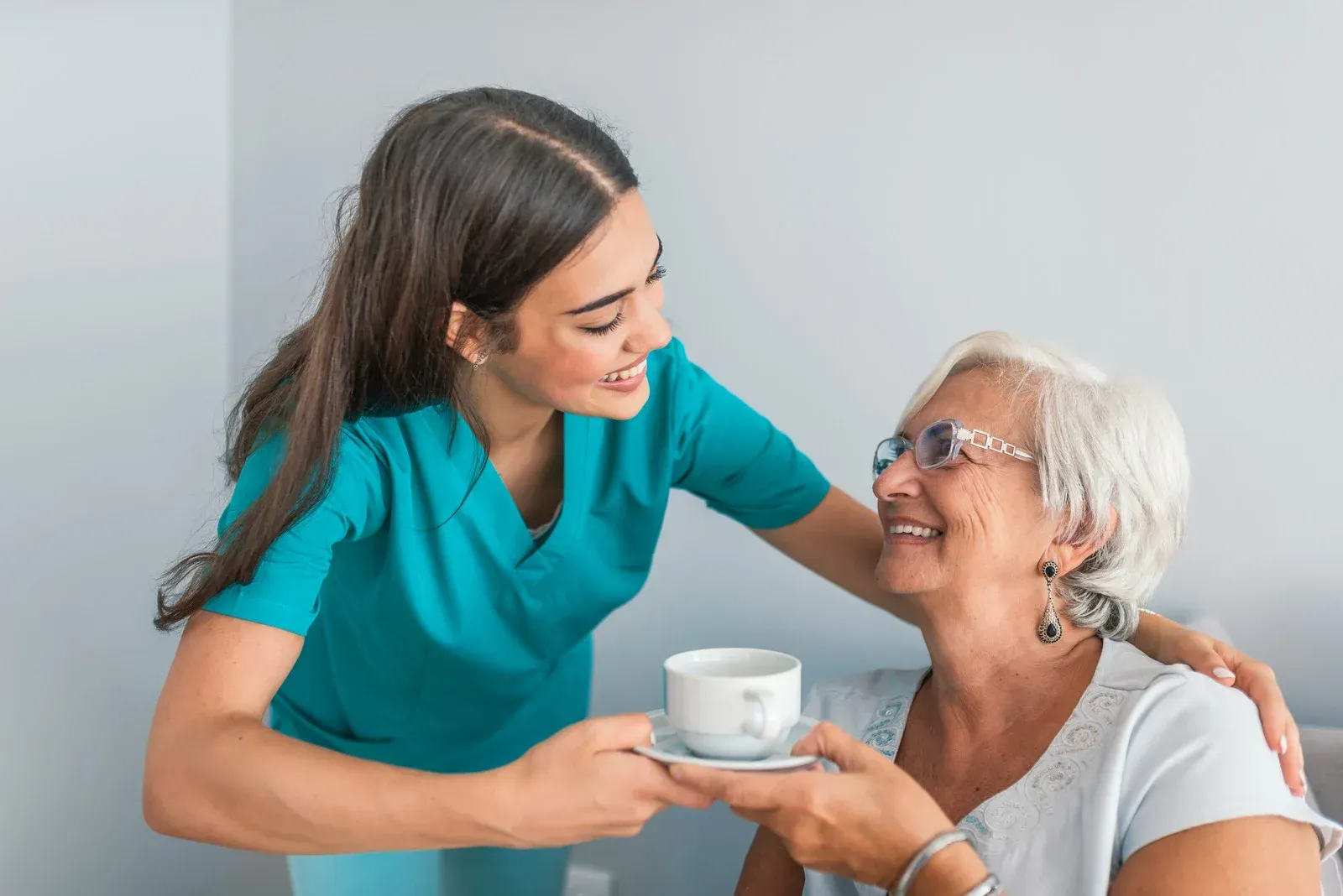 Caregiver offers a cup to an elderly person indoors, both smiling.