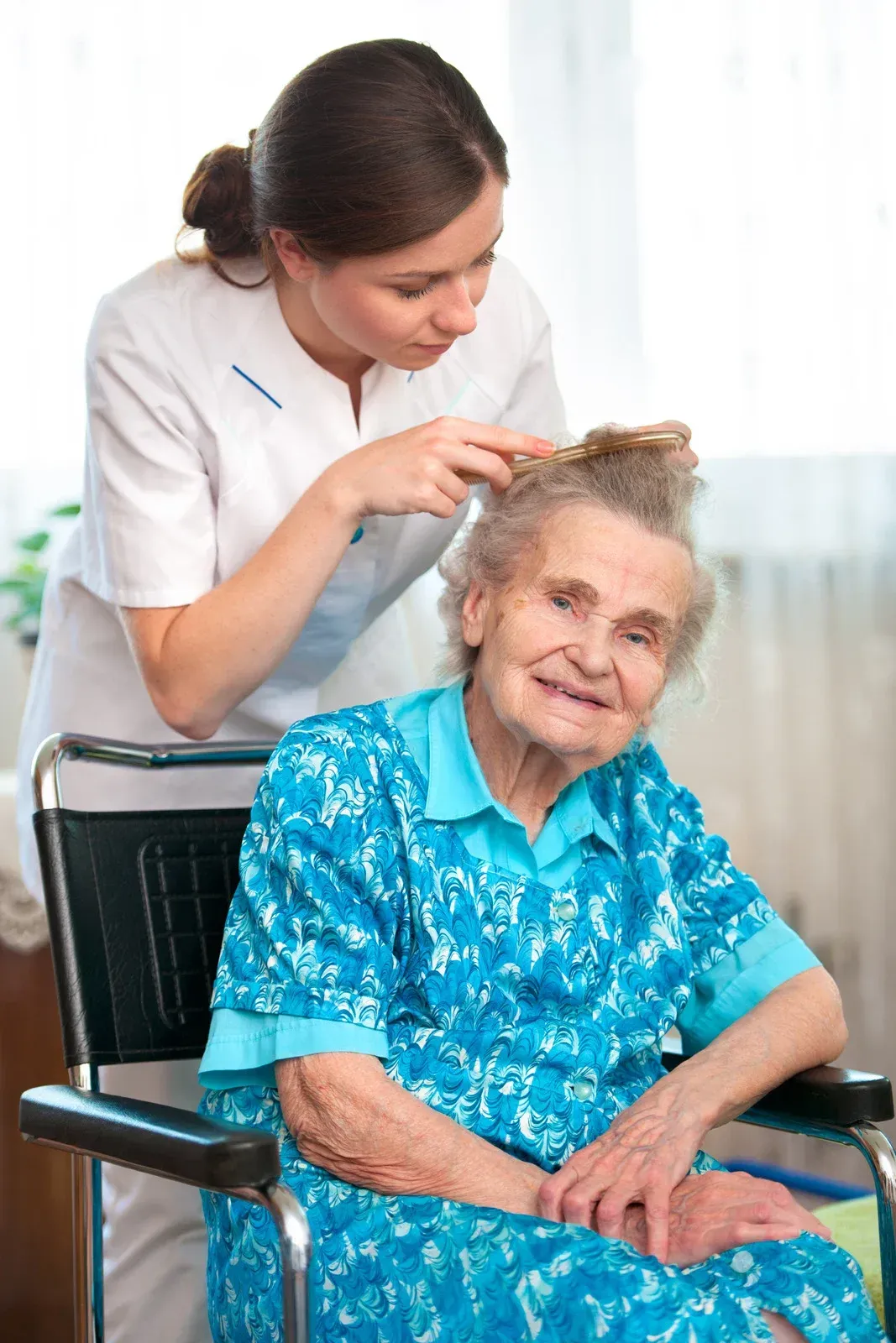 Caregiver brushing hair of elderly woman in wheelchair; indoor setting.