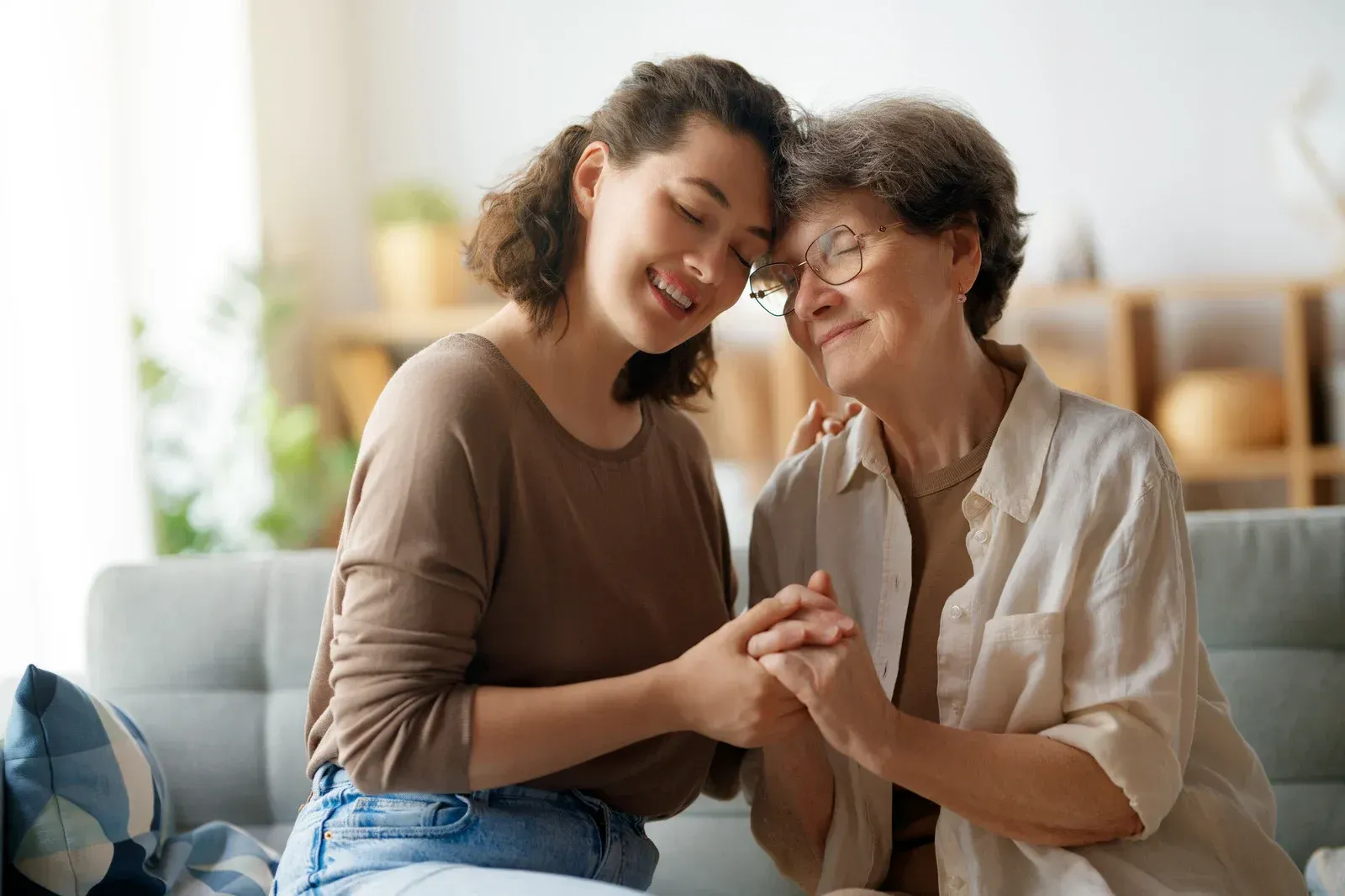 Woman and older person holding hands, smiling warmly, sitting on a couch.