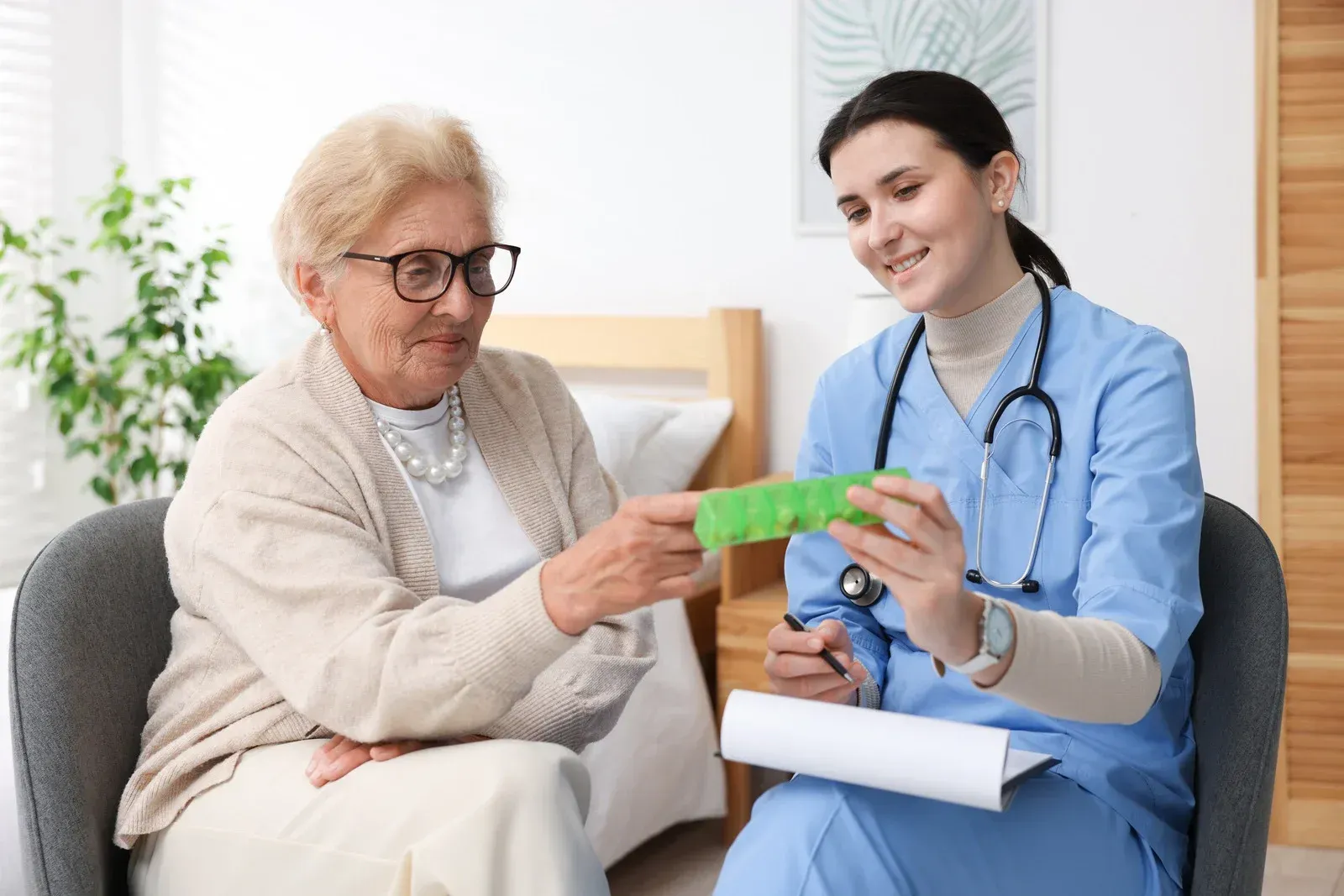 Nurse shows pill organizer to senior woman, bedroom setting.