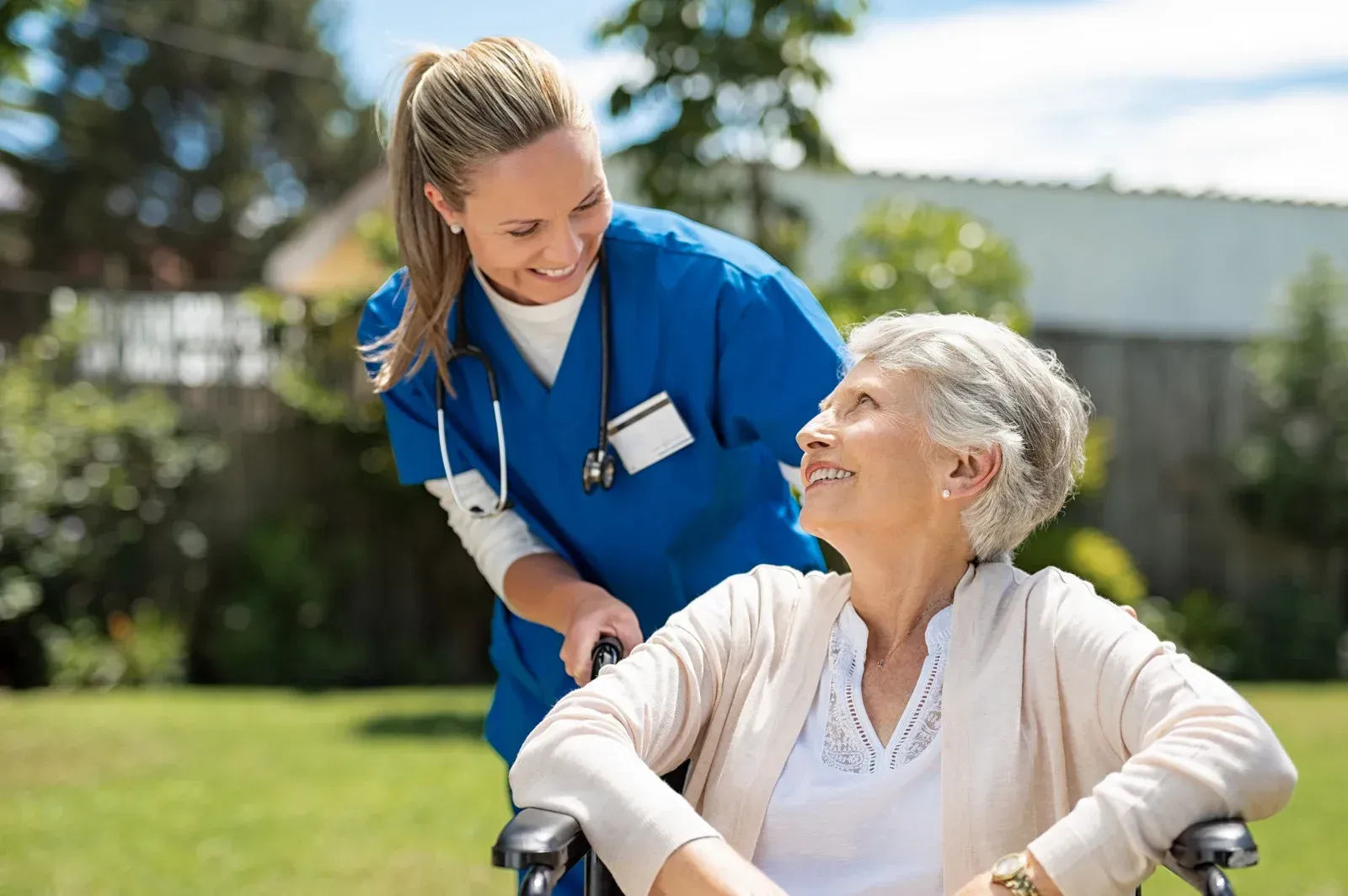 Nurse in blue scrubs smiles at woman in a wheelchair, outdoors.