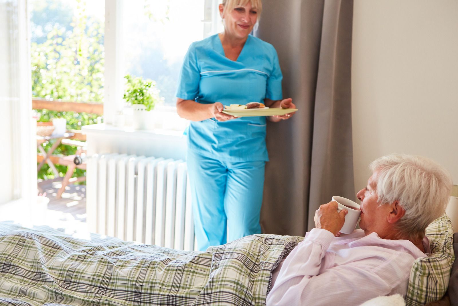 Nurse in blue scrubs delivers tray to a person drinking from a mug in bed near a window.