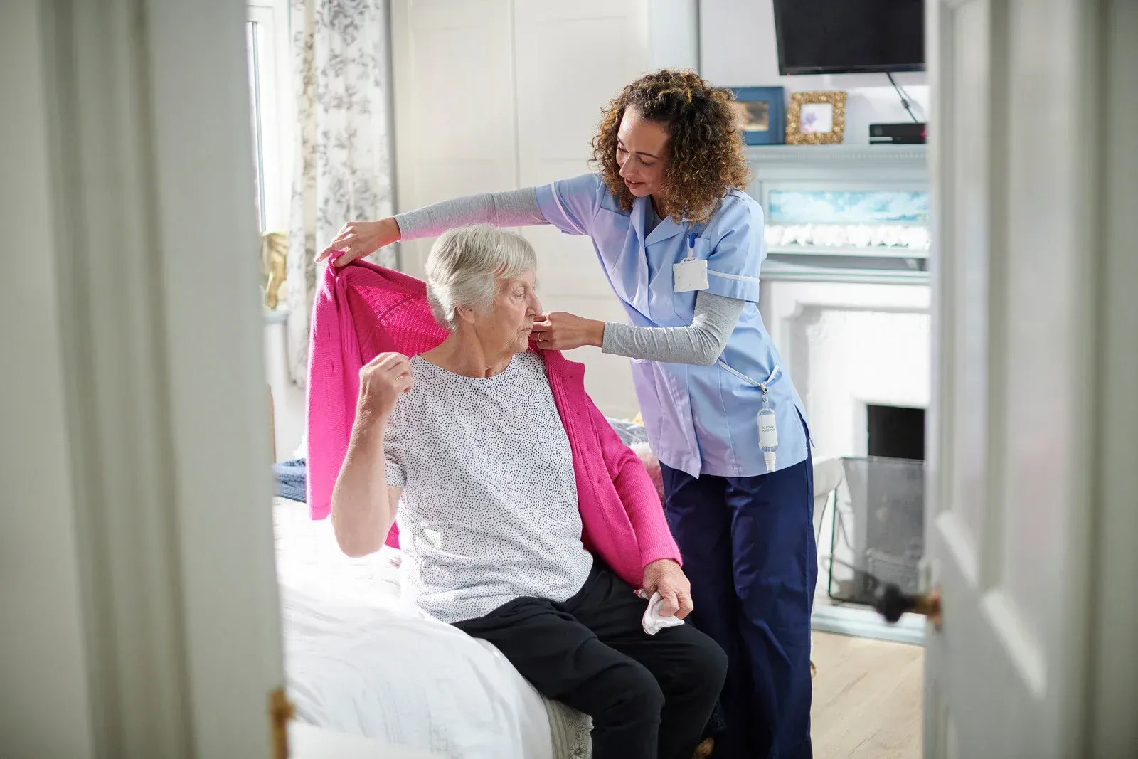 A caregiver helping an elderly person put on a pink sweater in a bedroom.