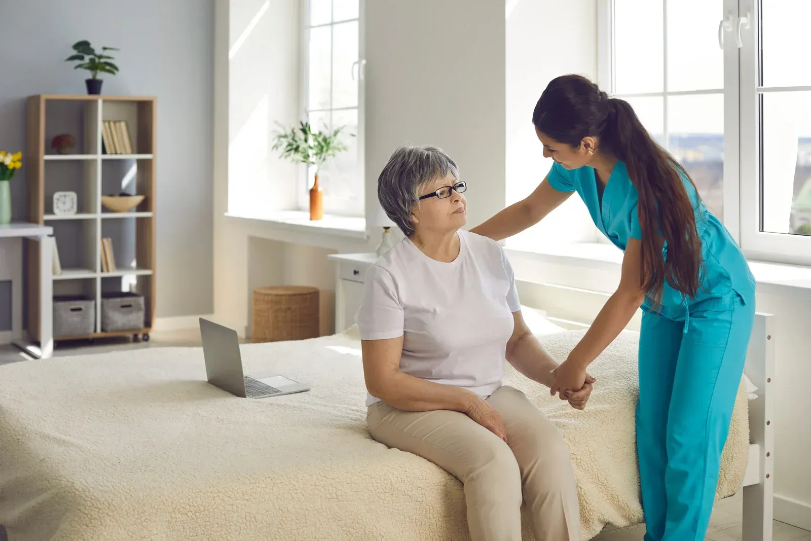 Caregiver assists a person sitting on a bed. Bright room with laptop and shelf.