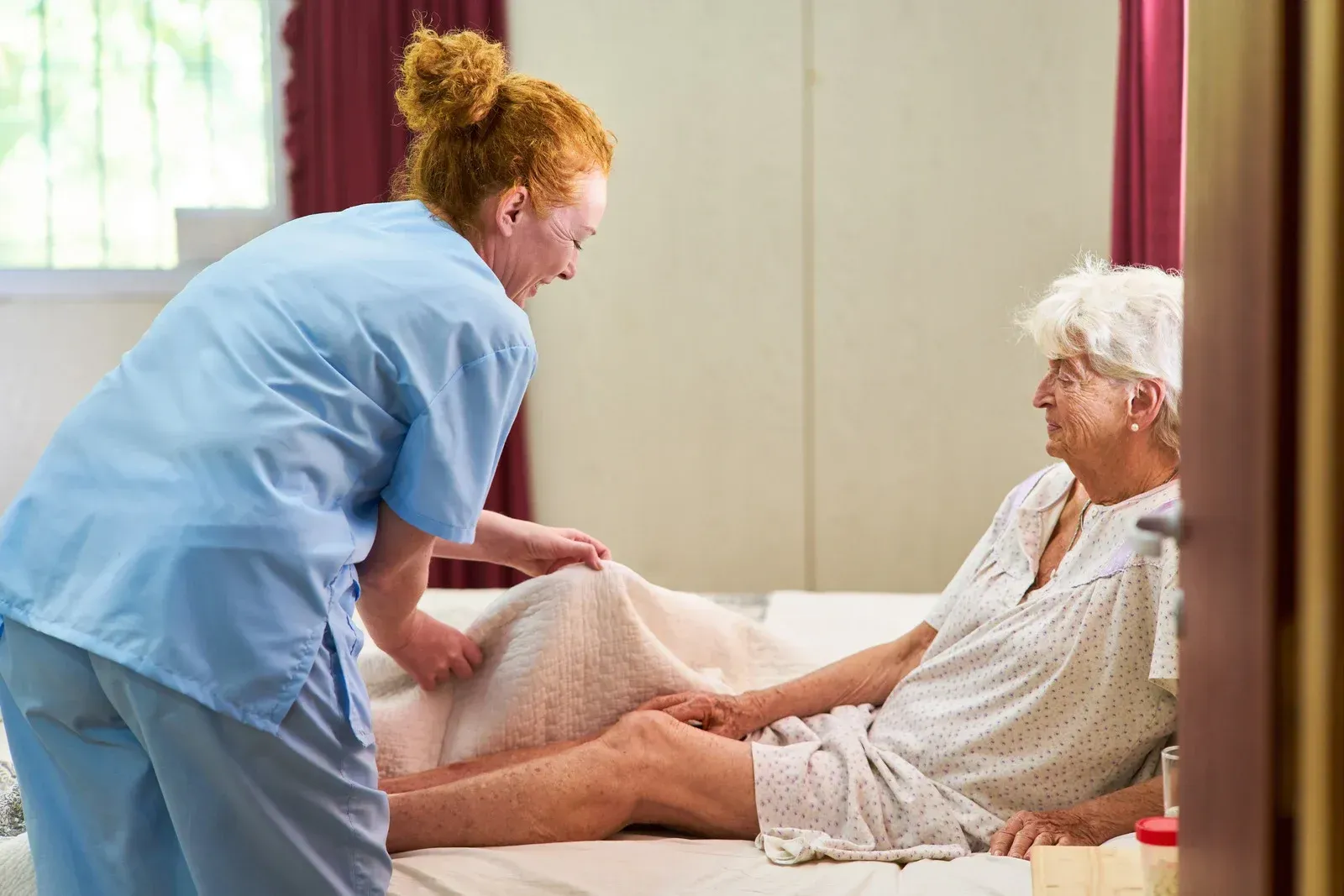 Caregiver assisting a person in bed, covering their legs with a blanket.