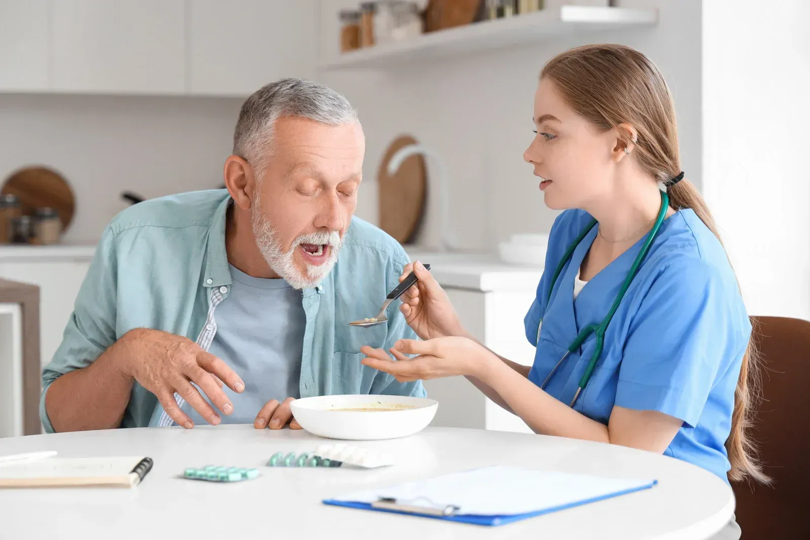 A caregiver feeds a man at a table, both in a kitchen. The man is opening his mouth.