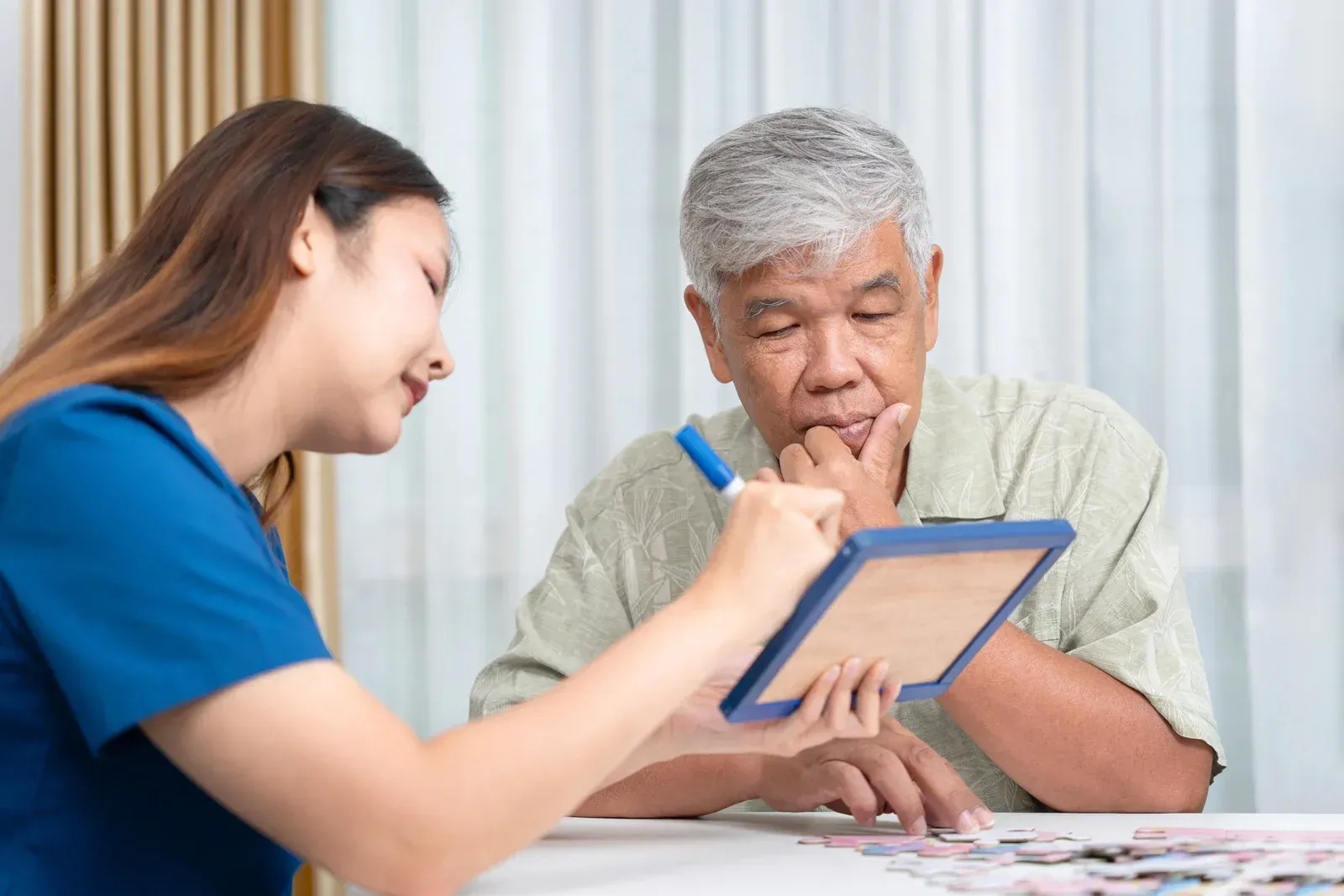 Woman in blue writes on a clipboard as older man looks on, at a table with puzzle pieces.