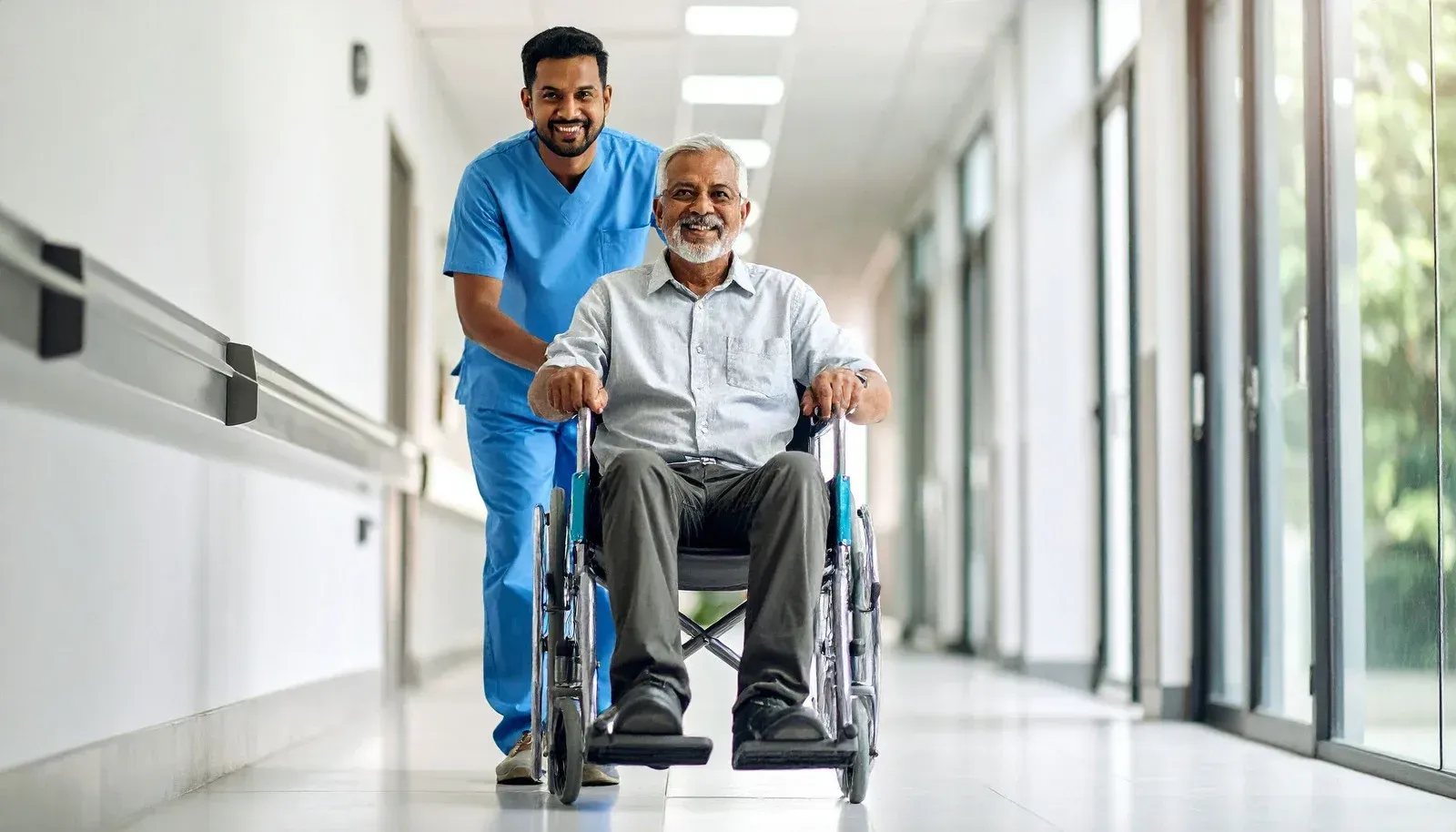 Healthcare worker pushing a person in a wheelchair down a sunlit hallway.