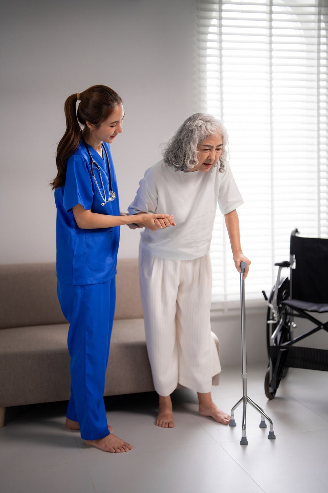 Nurse in blue scrubs smiles at senior man, holding clipboard, in a kitchen.