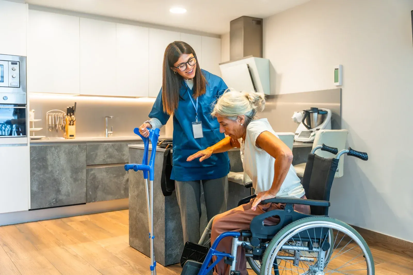 Caregiver assists person in wheelchair to stand in kitchen; crutch nearby.