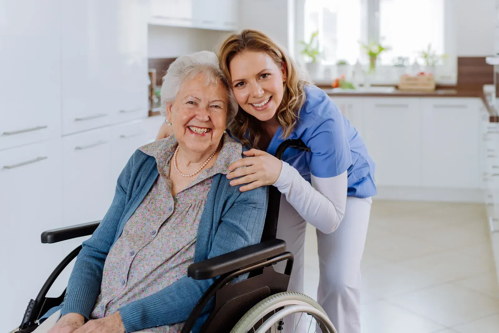 Woman in wheelchair smiles with caregiver, both smiling in a bright kitchen.