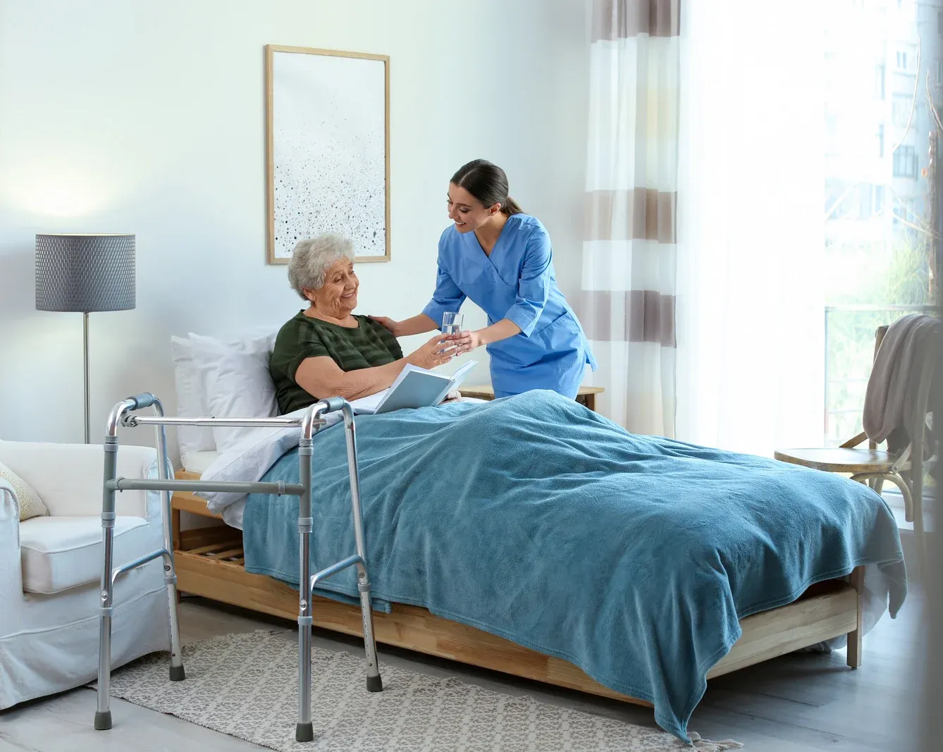 Nurse assisting patient in bed; blue scrubs, smiling; walker nearby; light room.