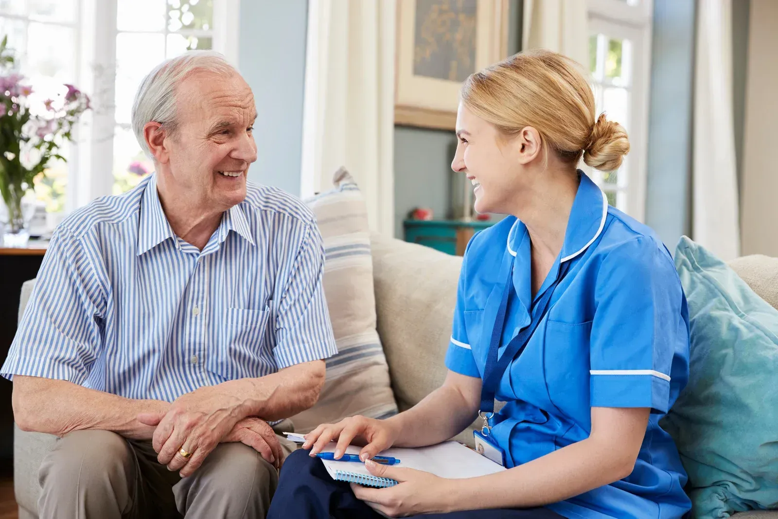 Elderly man and nurse in blue scrubs talking on a couch indoors.
