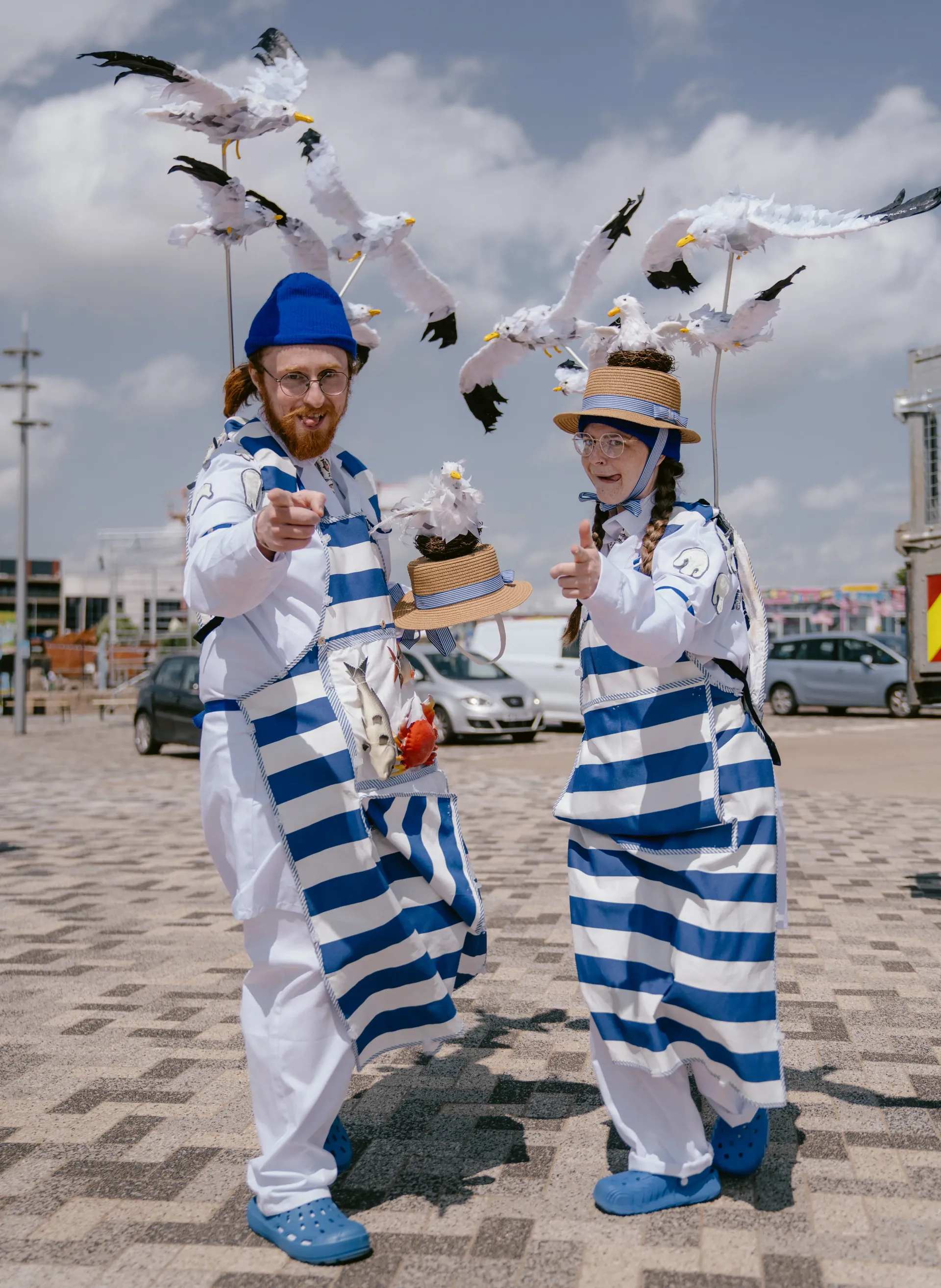Two performers in blue-and-white striped outfits stand outdoors on a sunny day, pointing toward the camera. They wear whimsical hats decorated with small nests and have tall poles attached to their costumes holding multiple seagull figures posed as if flying above them. Cars and buildings are visible in the background under a partly cloudy sky.